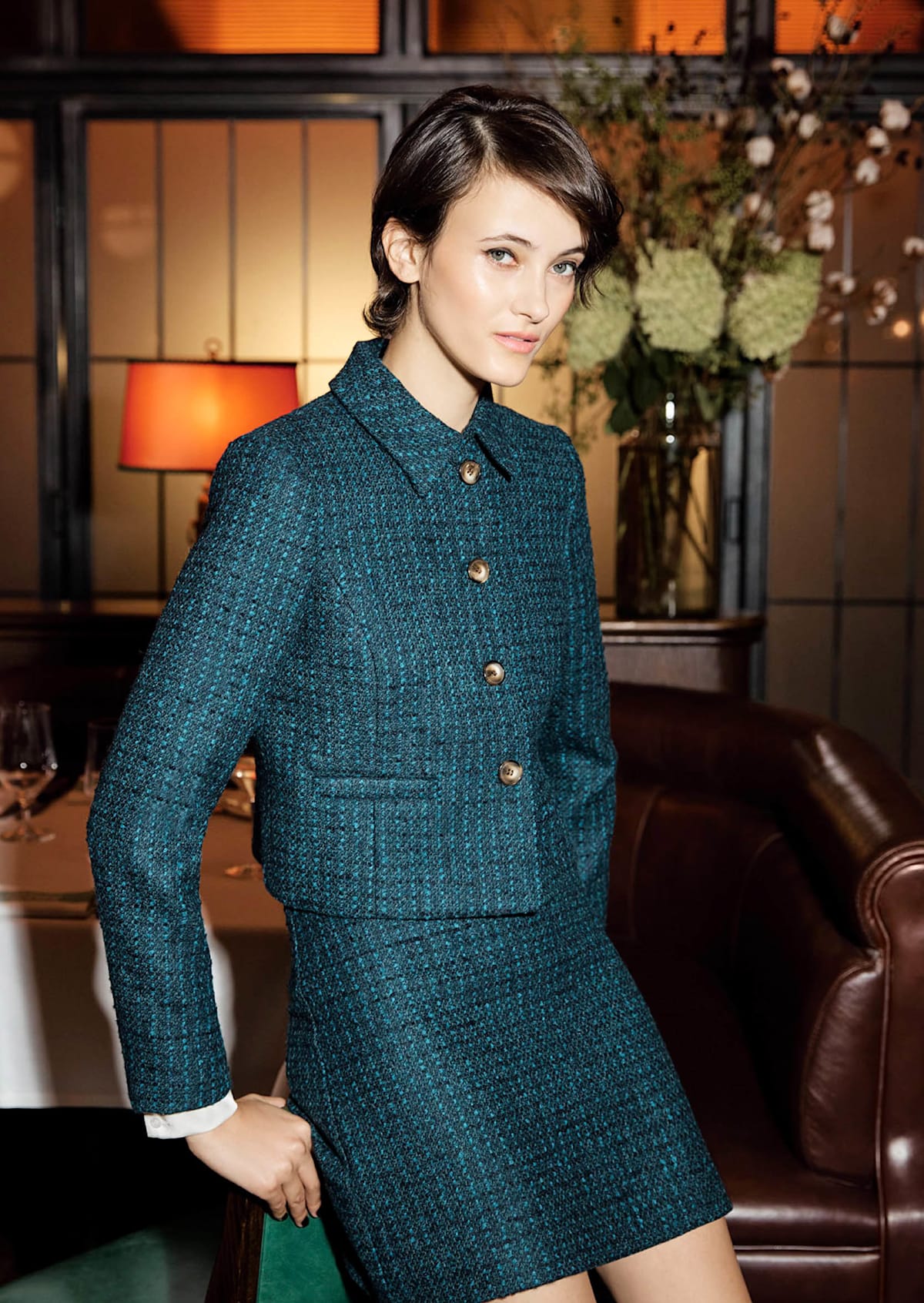 Model photographed sitting and standing in front of a chair wearing a satin shirt and a tweed jacket and skirt