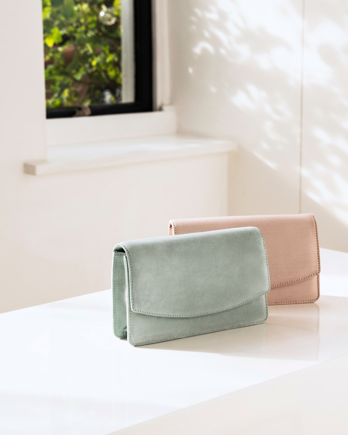 One mint green clutch bag and one oyster clutch bag photographed on a window ledge.